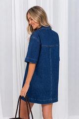 A woman wearing a dark blue denim shirt dress, shown from the back and side, holding a woven brown tote bag against a white curtain background.