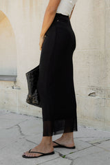 A person wearing a long black midi skirt with a sheer ankle hem, paired with a white top and black flip-flops, standing outdoors against a light stone wall.