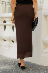 A woman wearing a long, dark brown satin midi skirt, paired with a black top, black heels, and holding a black clutch, standing outdoors in front of a white stone wall.