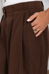 Close-up of a person wearing brown pleated wide-leg trousers, with a hand inserted into the side pocket.