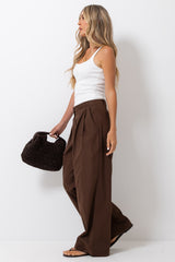 A woman stands in profile wearing a white tank top, brown wide-leg pleated pants, and holding a black woven handbag against a plain white studio background.