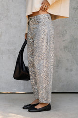 A person wearing wide-leg cropped pants with a beige and gray animal print pattern, styled with a cream knit top, black flat shoes, and holding a black leather shoulder bag against a gray concrete wall.