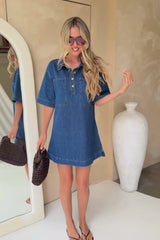 A woman modeling a short-sleeve blue denim shirt dress with a buttoned collar, paired with a dark brown woven handbag and brown flip-flop sandals, against a beige wall.