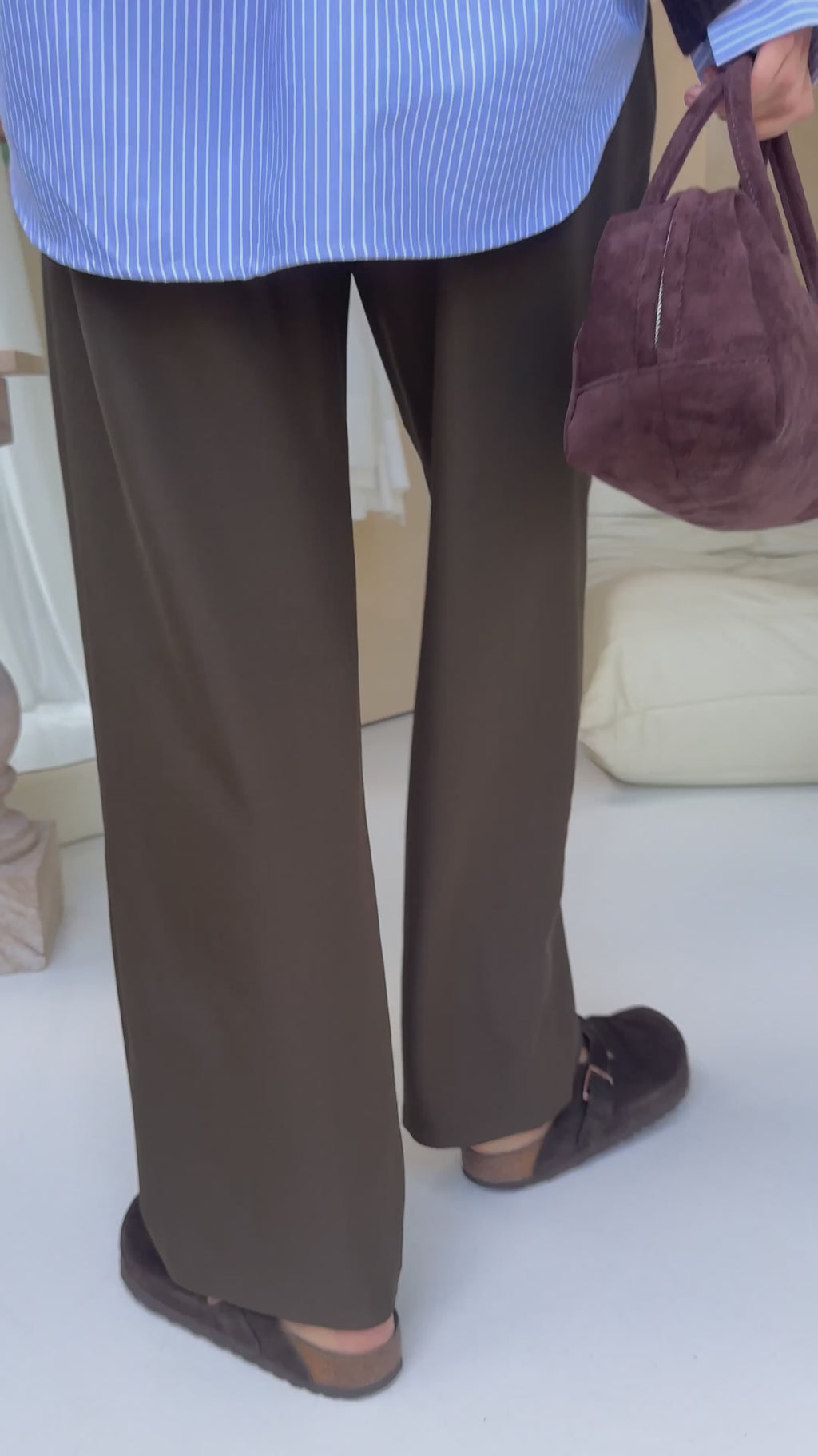 Xavier pants in dark olive brown styled with a brown oversized sweater, blue striped shirt, and brown suede clogs in a mirrored entryway setting