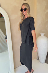 A woman modeling a black short-sleeve top and matching midi skirt set, styled with black sunglasses, a small dark woven handbag, and black strappy heels against a beige wall.