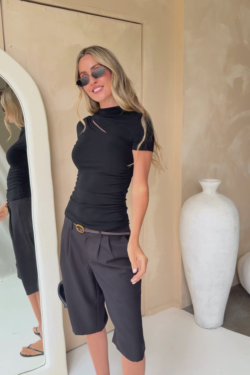 A woman modeling a black short-sleeve top with a diagonal cutout and black midi shorts, styled with a black belt, black sunglasses, and holding a black handbag against a beige wall studio background.
