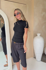 A woman modeling a black short-sleeve top with a diagonal cutout and black midi shorts, styled with a black belt, black sunglasses, and holding a black handbag against a beige wall studio background.