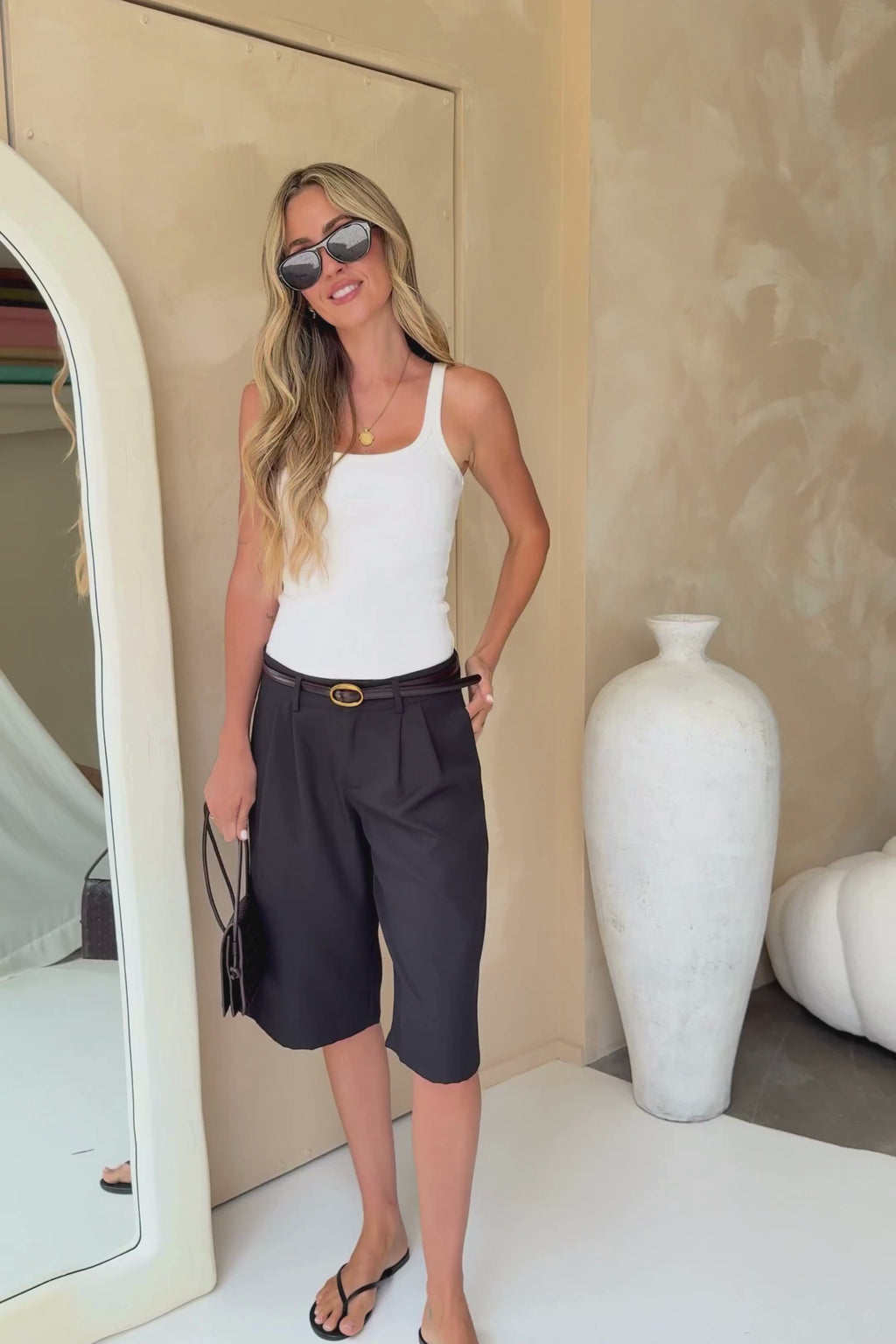 A woman modeling black pleated culotte shorts with a black belt, styled with a white tank top, black sunglasses, and a small black shoulder bag against a beige wall.