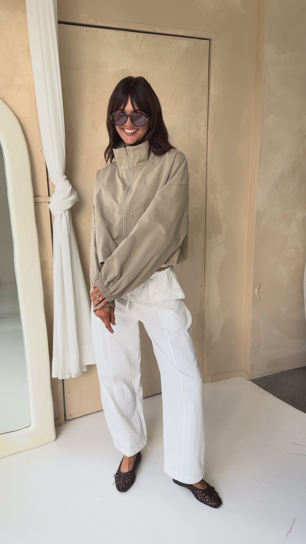 Woman wearing a stone funnel neck bomber jacket over white pants.