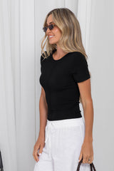 A woman wearing a fitted black short-sleeve top and white drawstring pants, standing against a light curtain background.
